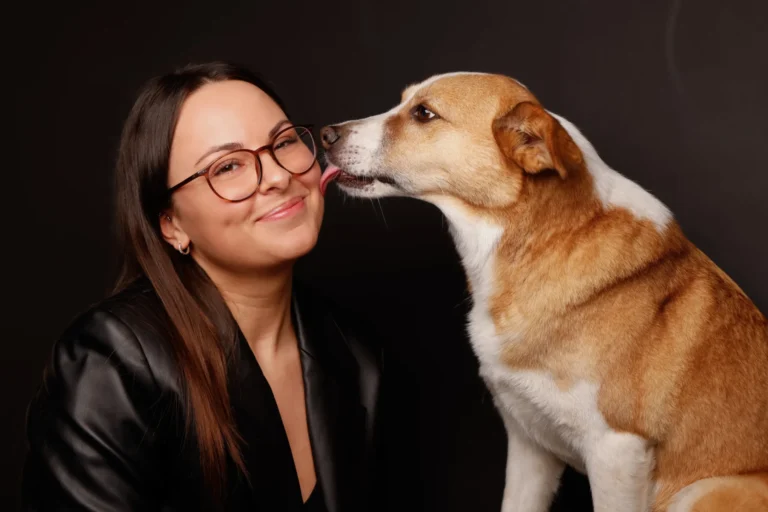 Eine lächelnde Frau mit Brille sitzt neben einem Hund, der sie mit der Zunge an der Wange leckt, vor dunklem Hintergrund.
