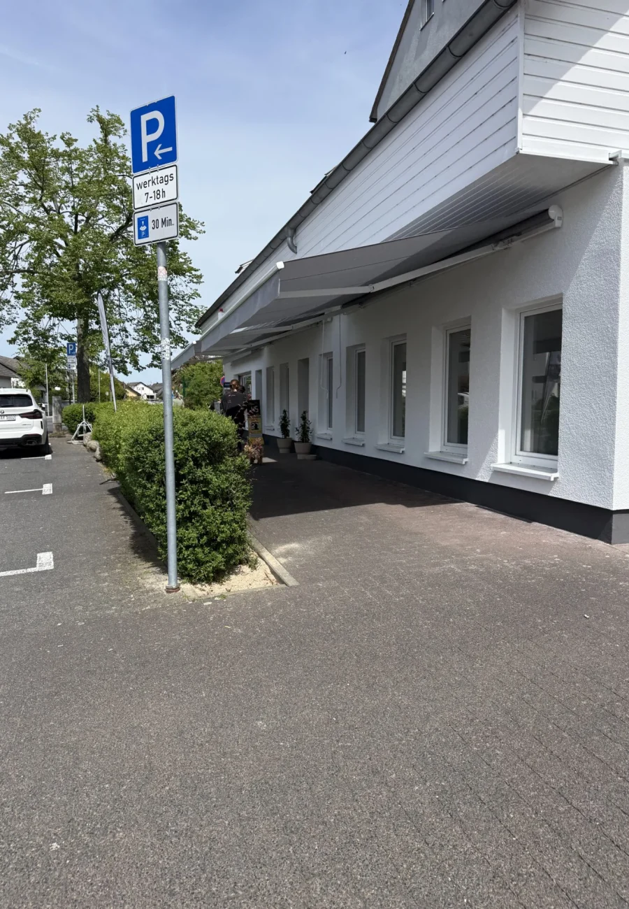 Parkplatz mit Schild für Parkzeiten, angrenzendes Gebäude mit weißen Wänden und grüner Bepflanzung.